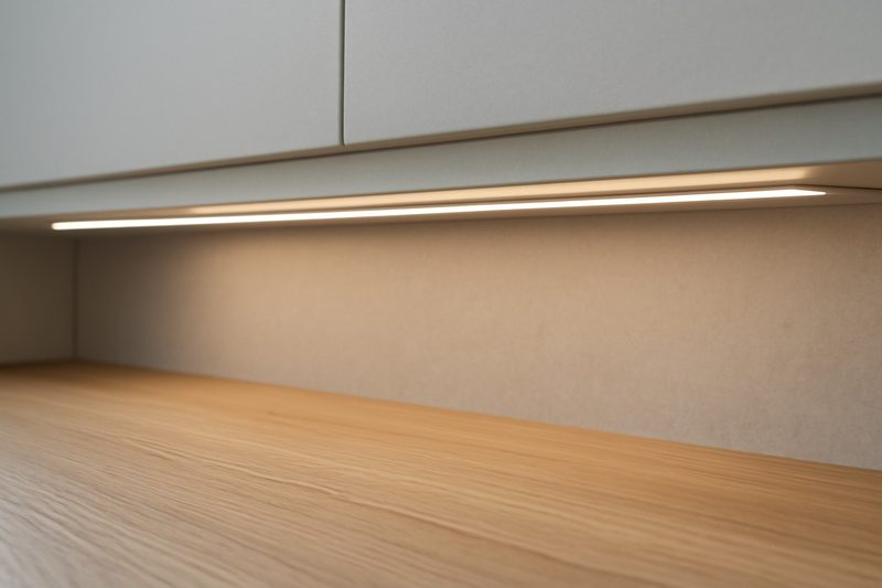 نمای نزدیک از نوار نازک LED نصب‌شده زیر لبهٔ کابینت که روی سطح کار چوبی نور می‌تاباند