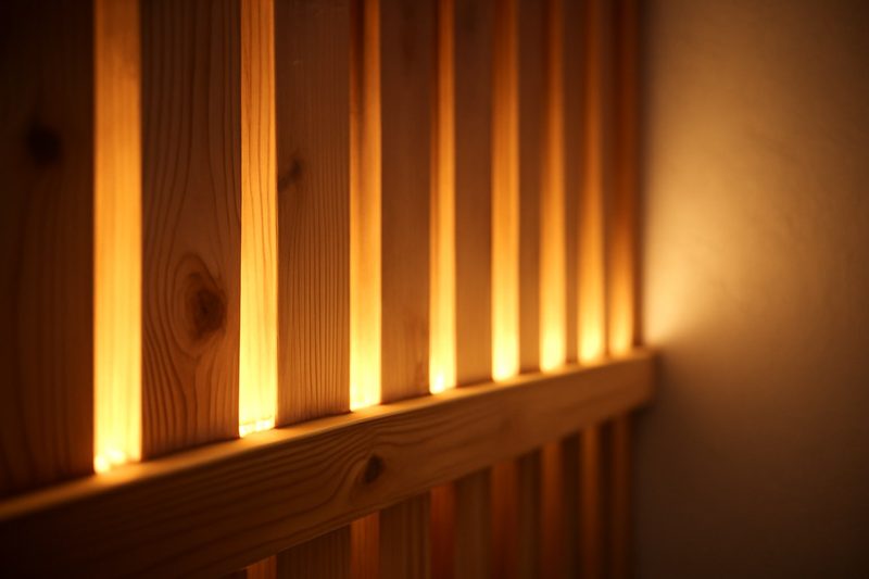 نمای نزدیک دیوارپوش چوبی مشبک با نوار LED گرم درخشان میان تخته‌ها